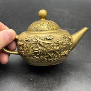 May include: A metallic gold-toned teapot featuring intricate embossed relief carvings of a phoenix bird surrounded by stylised clouds. The teapot has a rounded body, a curved spout, a small handle, and a matching lid with a spherical knob.