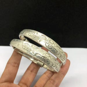Pode incluir: Dois braceletes de metal prateado com um padrão floral em relevo de flores de cinco pétalas e folhas curvas. As pulseiras têm um acabamento liso e polido, com um mecanismo de dobradiça em forma de mola visível na lateral das bandas.
