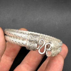 Könnte beinhalten: Ein silberfarbenes geflochtenes Armband mit einem zweisträngigen Kettenmuster. Das Armband wird mit einem S-förmigen Hakenverschluss geschlossen und verfügt über zwei zylindrische Endkappen, die mit komplexen Prägemustern und seilartigen Texturen verziert sind.