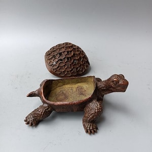 May include: A bronze-coloured metal turtle-shaped container with a removable lid. The lid features a textured pattern of overlapping circular coin motifs. The interior of the turtle body is hollow with a smooth, metallic finish, while the exterior has a dark, textured, weathered appearance.