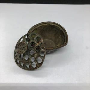 May include: A small, dark bronze-toned metal container with a separate lid. The lid features a circular, perforated design resembling a lotus seed pod with numerous open holes. The base is a simple, open bowl shape with a weathered, metallic finish.