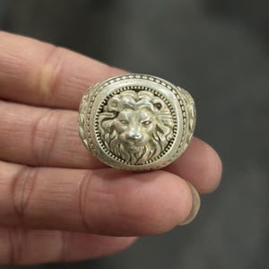 Puede incluir: Un anillo de metal plateado con una cara circular que presenta un relieve detallado de una cabeza de león. La banda del anillo y el borde que rodea al león están decorados con un patrón de cuentas texturizado, dándole un acabado metálico.