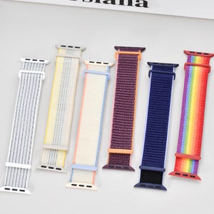 Puede incluir: Una colección de seis correas de reloj en varios colores. Las correas incluyen gris, amarillo, blanco con azul, morado, azul marino y un patrón de arcoíris. Cada correa tiene una textura tejida y está diseñada para un reloj inteligente.