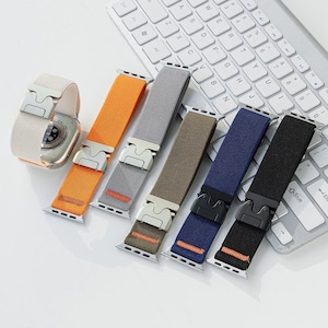 Puede incluir: Una colección de correas para smartwatch en varios colores: naranja, gris, verde oliva, azul marino y negro. Cada correa tiene un cierre metálico y está diseñada para un smartwatch. Las correas se muestran cerca de un teclado blanco.