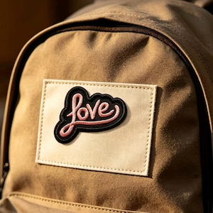 May include: A tan backpack featuring a rectangular cream-colored patch on the front. Centered on the patch is an embroidered black and pink emblem with the word love written in a cursive script style, featuring a thick black border outline.