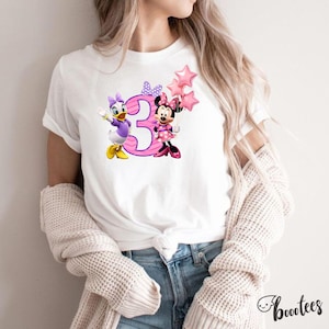 May include: A white short-sleeved t-shirt featuring a graphic print of Daisy Duck and Minnie Mouse standing next to a large pink number three. Minnie Mouse holds a cluster of three pink star-shaped balloons, and both characters wear bows.