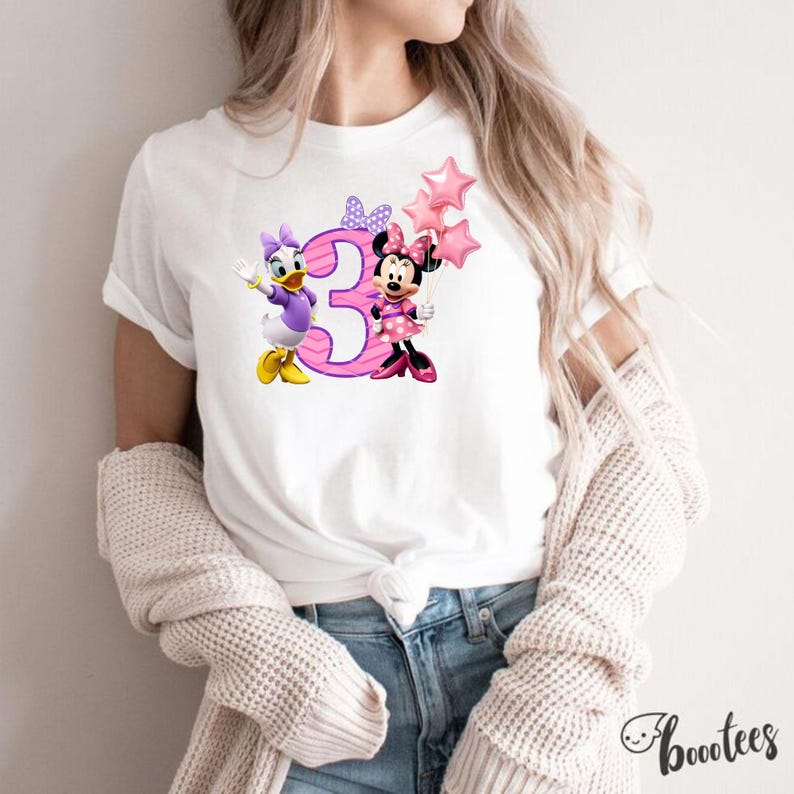 May include: A white short-sleeved t-shirt featuring a graphic print of Daisy Duck and Minnie Mouse standing next to a large pink number three. Minnie Mouse holds a cluster of three pink star-shaped balloons, and both characters wear bows.