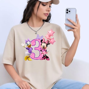 May include: A beige short-sleeved t-shirt featuring a graphic print of Daisy Duck and Minnie Mouse standing next to a large pink number three. Minnie Mouse holds three pink star-shaped balloons, and a purple polka-dot bow sits above the number.