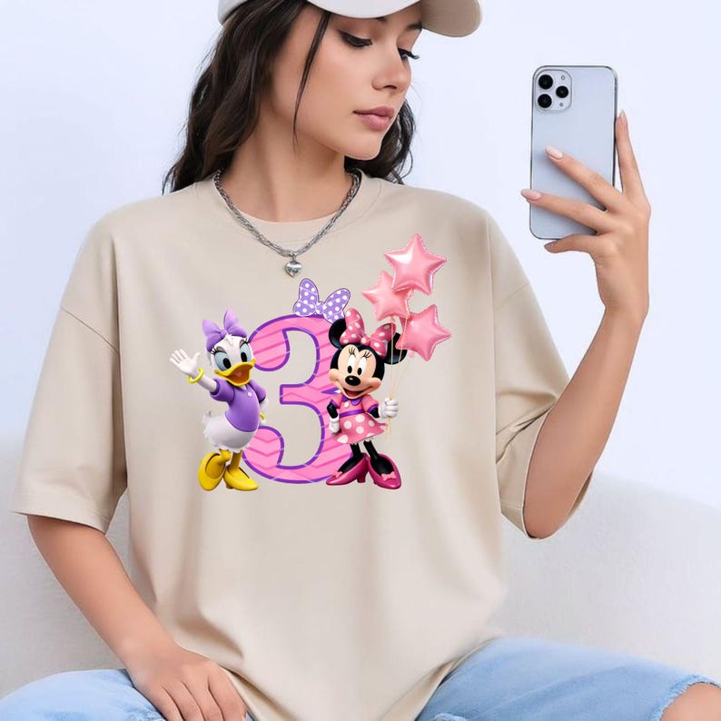 May include: A beige short-sleeved t-shirt featuring a graphic print of Daisy Duck and Minnie Mouse standing next to a large pink number three. Minnie Mouse holds three pink star-shaped balloons, and a purple polka-dot bow sits above the number.