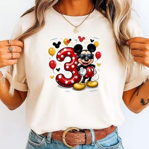 May include: A cream-colored t-shirt featuring a graphic of Mickey Mouse wearing sunglasses next to a large red number three with white polka dots. The design includes yellow and red balloons and small black Mickey Mouse head silhouettes.