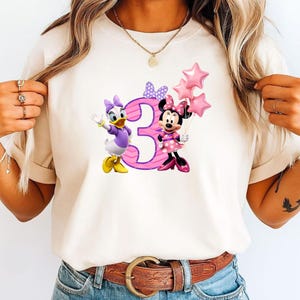 May include: A cream-colored t-shirt featuring a graphic of Daisy Duck and Minnie Mouse standing next to a large pink number three. Minnie Mouse holds three pink star-shaped balloons. A purple polka-dot bow is positioned above the number three.