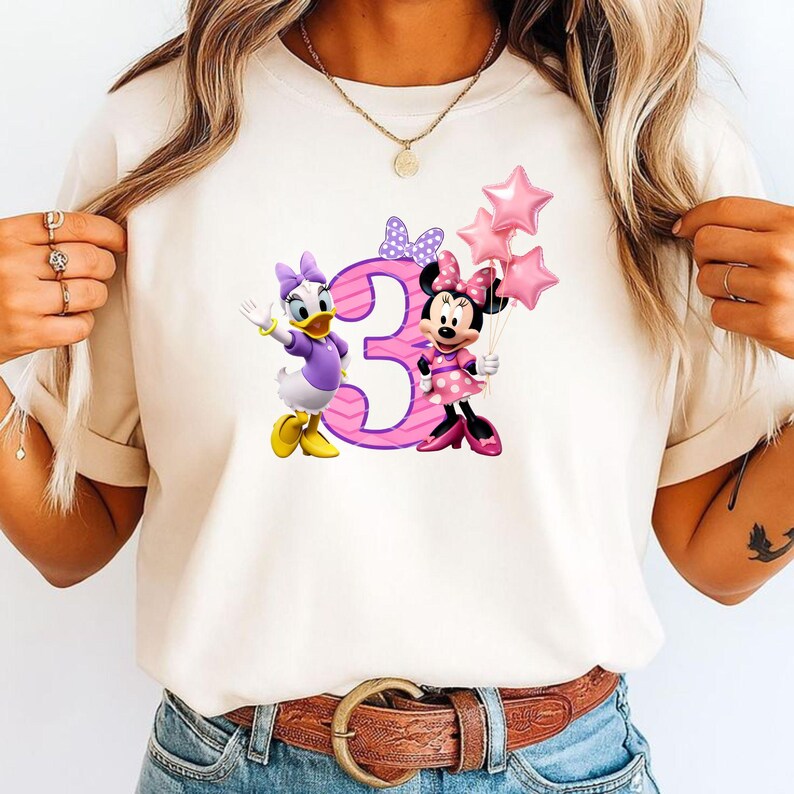 May include: A cream-colored t-shirt featuring a graphic of Daisy Duck and Minnie Mouse standing next to a large pink number three. Minnie Mouse holds three pink star-shaped balloons. A purple polka-dot bow is positioned above the number three.