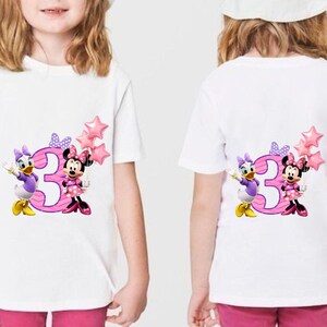 May include: A white short-sleeved t-shirt featuring a graphic of the number three in pink with purple stripes, accompanied by Daisy Duck and Minnie Mouse. Minnie holds a cluster of three pink star-shaped balloons, and both characters wear bows.