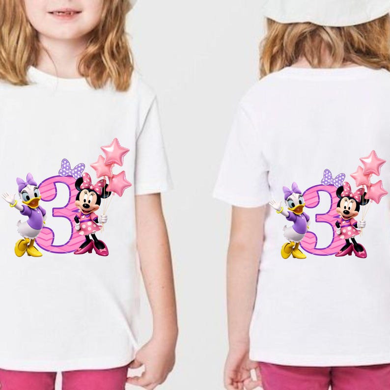 May include: A white short-sleeved t-shirt featuring a graphic of the number three in pink with purple stripes, accompanied by Daisy Duck and Minnie Mouse. Minnie holds a cluster of three pink star-shaped balloons, and both characters wear bows.