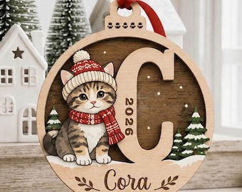 May include: A round wooden Christmas ornament featuring a layered design with a tabby kitten wearing a red and white knit hat and scarf. The ornament displays a large letter C, the year 2026, and the name Cora, accented by a small red heart.