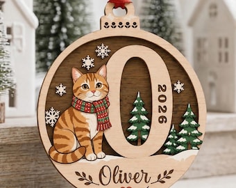 May include: A round wooden Christmas ornament featuring a ginger tabby cat wearing a red and green scarf, the letter O, the year 2026, and the name Oliver. The design includes small green pine trees, white snowflakes, and a red ribbon hanger.