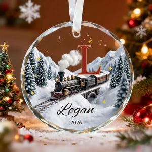 Puede incluir: Un adorno navideño circular de cristal que muestra un tren de vapor negro viajando a través de un paisaje montañoso nevado con pinos. El diseño incluye una letra L roja grande, el nombre Logan en cursiva negra y el año 2026 en negro.