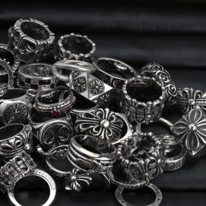 Könnte beinhalten: Eine Sammlung von silberfarbenen Metallringen mit komplexen Prägemustern, darunter florale Motive, Kreuzmuster und geometrische Formen. Einige Ringe zeigen kleine eingebettete Steine, während andere detaillierte Gravuren und strukturierte Oberflächen aufweisen.