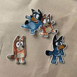 May include: Four embroidered patches featuring cartoon dog characters. One patch shows two dogs jumping together, while two separate patches depict individual dogs in orange and blue colors with white accents and black outlines.
