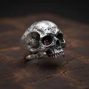 May include: A silver ring shaped like a detailed human skull with a textured, pitted surface. The skull features a single small, round, faceted red gemstone set into the left eye socket, contrasting with the dark, oxidized silver finish.