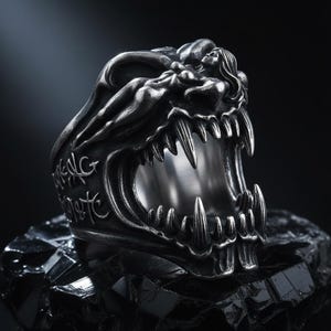 May include: A silver-toned metal ring featuring a sculpted creature with an open mouth displaying sharp, prominent teeth. The band is detailed with engraved markings and a figure with long hair integrated into the upper portion of the design.