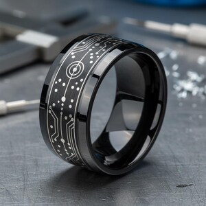 Könnte beinhalten: Ein schwarzer Ring mit polierter Oberfläche und einem lasergravierten weißen Leiterplattenmuster, das sich um das äußere Band zieht. Das Design umfasst feine Linien und kreisförmige Knoten, die das Aussehen elektronischer Schaltkreise nachahmen.