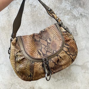 Puede incluir: Un bolso de mano marrón y beige con estampado de piel de serpiente y forma curvada. El bolso tiene cierre de cremallera, correa para el hombro y herrajes plateados. El diseño del bolso incluye una solapa y un fondo redondeado.