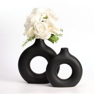 May include: Two matte black ceramic vases with a circular, donut-like shape and a central opening. The larger vase on the left contains a bouquet of white artificial roses with green leaves and stems, while the smaller vase on the right is empty.