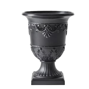 May include: A matte black urn planter featuring a wide flared rim, a tapered body with embossed scrollwork and leaf motifs, and a fluted pedestal base that rests on a square foundation. The vessel has a smooth finish and symmetrical design.