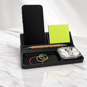 Op de afbeelding: Een zwarte bureau-organizer met een verticale gleuf voor een smartphone, een vak voor limoengroene memoblaadjes, een groef voor een pen met kurken afwerking en twee open vakken met kleurrijke elastiekjes en oordopjes.