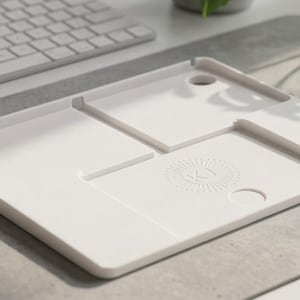 Peut inclure: Un plateau organisateur de bureau rectangulaire blanc doté de plusieurs compartiments encastrés de tailles et de formes variées, incluant des découpes circulaires et une section centrale avec un logo circulaire subtilement gaufré.