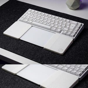 Puede incluir: Un teclado mecánico blanco con distribución QWERTZ, que cuenta con un panel táctil central flanqueado por dos reposamuñecas blancos. El reposamuñecas derecho muestra un logotipo circular con las letras KJ. El dispositivo está sobre una alfombrilla gris.