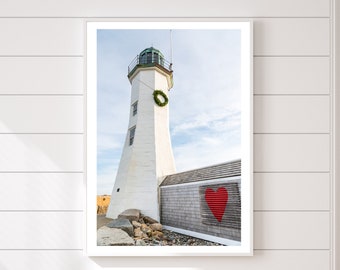 May include: A white lighthouse with a green roof and a red heart painted on the wall behind it. The lighthouse is on a rocky shore with a blue sky in the background.