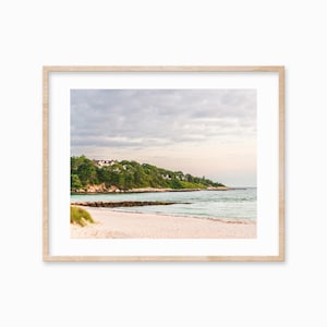 May include: A scenic photograph of a sandy beach with a rocky shoreline and a line of trees and houses in the distance. The sky is cloudy and the water is a light blue color.
