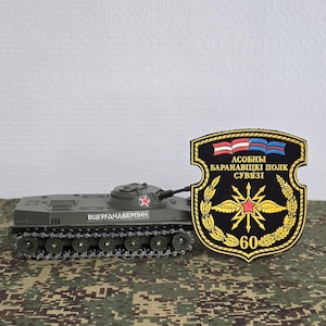 Puede incluir: Un tanque de juguete verde oliva con una estrella roja y texto cirílico en el lateral, junto a un parche militar negro en forma de escudo con un emblema dorado, una estrella roja y el texto ASOBNY BARANAVITSKI POLK SUVYAZI 60.