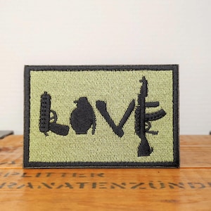 Peut inclure: Un écusson rectangulaire brodé sur fond vert olive clair avec une bordure noire. La broderie noire représente le mot LOVE, où la lettre L est une arme de poing, le O est une grenade, le V est formé par deux couteaux croisés et le E est un fusil.