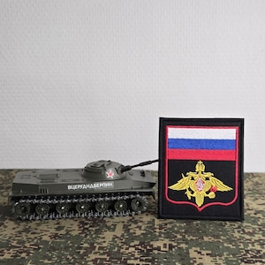 Könnte beinhalten: Ein olivgrüner Spielzeugpanzer mit einem roten Stern und kyrillischem Text an der Seite, platziert neben einem rechteckigen gestickten Aufnäher mit einer horizontalen weiß-blau-roten Flagge über einem goldenen Doppeladler-Emblem auf schwarzem Grund.