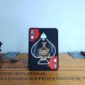 Puede incluir: Un parche rectangular negro bordado con un diseño de carta de as de picas. El centro del símbolo de pica contiene un gráfico detallado de una calavera. Aparecen acentos rojos detrás de los símbolos de las cartas en la esquina superior izquierda e inferior derecha.