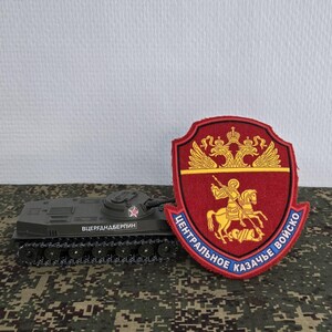 Pode incluir: Um tanque de brinquedo verde-escuro com uma pequena estrela vermelha na torre e texto cirílico na lateral, posicionado ao lado de um emblema bordado em forma de escudo com uma águia bicéfala dourada e um cavaleiro a cavalo em vermelho e ouro.