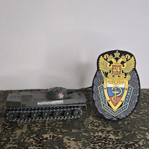 Puede incluir: Un modelo de tanque gris con una estrella roja en la torreta y el texto cirílico "ВЦЕРГАНДБЕРПИН" en el lateral, conectado por un cable fino a un parche bordado en forma de escudo con un águila bicéfala dorada y un emblema médico azul.