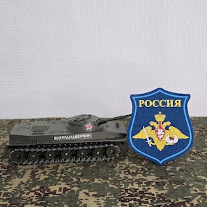 Könnte beinhalten: Ein olivgrüner Panzermodellbausatz mit einem roten Stern auf dem Turm und kyrillischem Text an der Seite, platziert neben einem blauen schildförmigen Aufnäher mit einem goldenen Doppeladler-Emblem und dem russischen Wort РОССИЯ in Gelb.