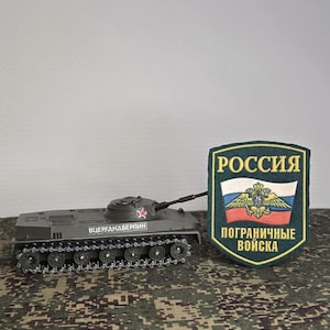 May include: A gray scale model tank featuring a red star on the turret and Cyrillic text on the side, positioned next to a dark green shield-shaped patch with the Russian flag, a golden eagle emblem, and the Cyrillic text ROSSIYA POGRANICHNYE VOYSKA.