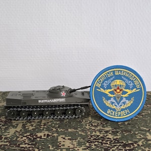 Op de afbeelding: Een grijs modeltankje met een kleine rode ster op de koepel en cyrillische tekst op de zijkant, naast een ronde blauwe geborduurde patch met een gele parachute, een rode ster en cyrillische tekst in een cirkelvormige rand.
