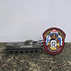 Puede incluir: Un tanque de juguete verde oliva con una estrella roja y texto cirílico en el lateral, colocado junto a un parche bordado en forma de escudo que muestra un oso, un paracaídas, un ancla y texto cirílico con un esquema de color rojo, blanco y azul.