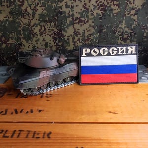 Peut inclure: Un modèle de char miniature camouflage placé à côté d'un écusson rectangulaire arborant le drapeau russe et le texte cyrillique РОССИЯ en lettres blanches sur fond noir, avec des bandes horizontales blanche, bleue et rouge en dessous.