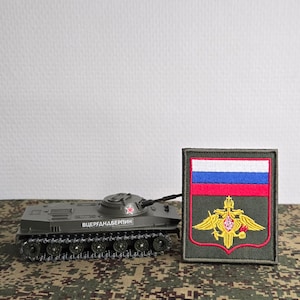 Op de afbeelding: Een miniatuur olijfgroen militair tankmodel met een rode ster op de koepel en cyrillische tekst aan de zijkant, geplaatst naast een rechthoekige geborduurde patch met de Russische vlag en een gouden dubbelkoppig adelaarsembleem.