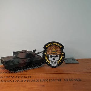Peut inclure: Un char d'assaut miniature noir placé à côté d'un écusson brodé circulaire représentant un crâne blanc portant un casque beige avec un petit drapeau bleu et jaune, entouré de texte et de rayons jaunes sur un fond noir à bordure cousue.