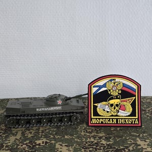May include: An olive green toy tank with a red star and Cyrillic text on the side, positioned next to a semi-circular embroidered patch featuring a skull, a dagger, a golden eagle emblem, and the Cyrillic text MOPCKAЯ ПEXOTA in yellow lettering.