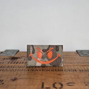 Può includere: Una toppa rettangolare con un motivo mimetico nei toni del marrone, verde e nero. Al centro della toppa si trova una grafica arancione brillante di una faccina sorridente stilizzata e maliziosa con occhi a punta e un ampio sorriso curvo.
