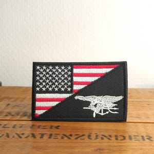 Peut inclure: Un écusson rectangulaire brodé présentant un design divisé. La partie supérieure gauche affiche le drapeau américain avec des couleurs rouge, blanc et bleu. La section inférieure droite est noire et arbore un emblème trident Navy SEAL brodé en blanc.